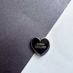 May include: A heart-shaped enamel pin with a gold border and the text "DORIAN HAVILLIARD" in gold lettering. The pin has a black center and a blue glitter accent along the bottom edge. The background is a diagonal split of black and white.
