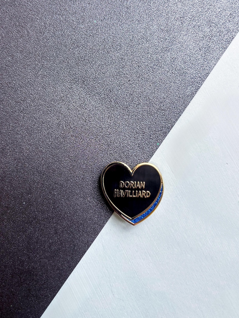 May include: A heart-shaped enamel pin with a gold border and the text "DORIAN HAVILLIARD" in gold lettering. The pin has a black center and a blue glitter accent along the bottom edge. The background is a diagonal split of black and white.