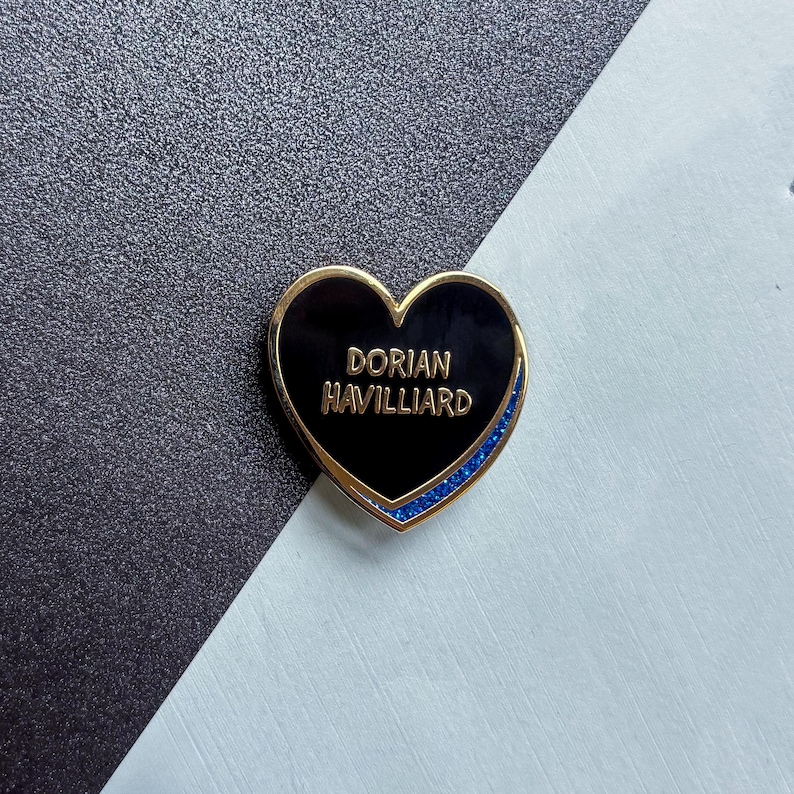 May include: A heart-shaped enamel pin with a black center and gold trim. The pin features the name "DORIAN HAVILLIARD" in gold lettering. A blue glitter border accents the heart's edge. The pin is set against a black and white background.