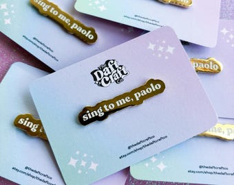 Könnte beinhalten: Goldene Emaille-Anstecknadel mit dem Text "sing to me, paolo" auf weißem Hintergrund. Die Anstecknadel befindet sich auf einer Karte mit dem Text "The Daft Craft Co." und Social-Media-Handles.