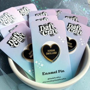 May include: A collection of enamel pins in a white bowl. Each pin is heart-shaped with a black center, gold trim, and the name "Dorian Havilliard." The pins are on cards with the text "The Daft Craft Co." and "Enamel Pin."