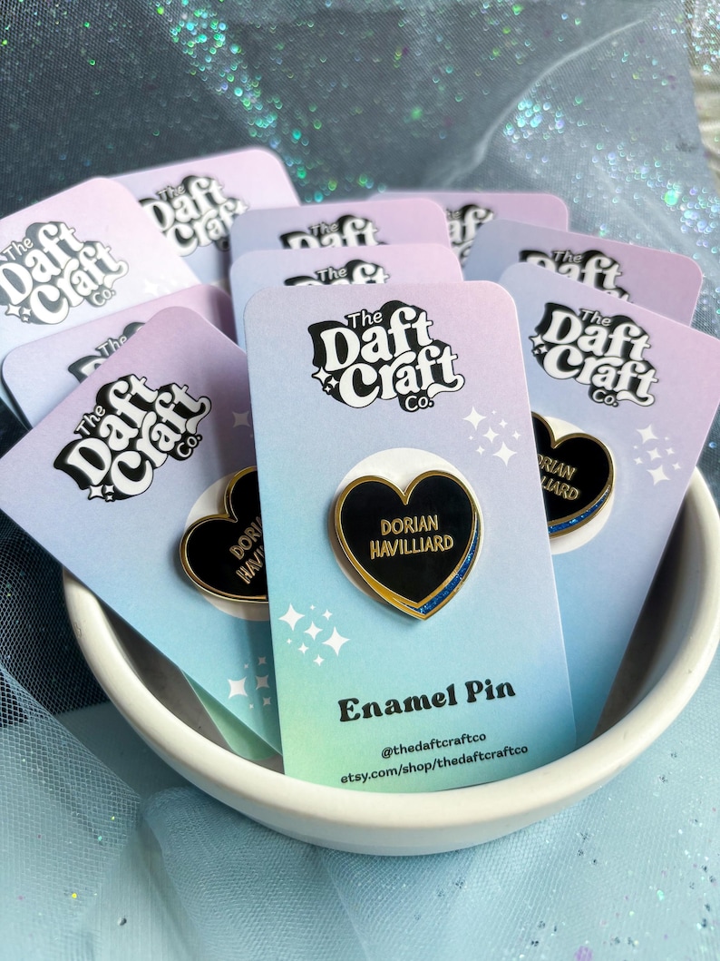 May include: A collection of enamel pins in a white bowl. Each pin is heart-shaped with a black center, gold trim, and the name "Dorian Havilliard." The pins are on cards with the text "The Daft Craft Co." and "Enamel Pin."