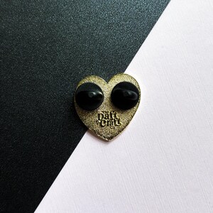 May include: A gold-colored heart-shaped enamel pin with two black pin backs. The pin has the text "Daft Craft Co." in a stylized font. The background is split diagonally, with black and white sections.