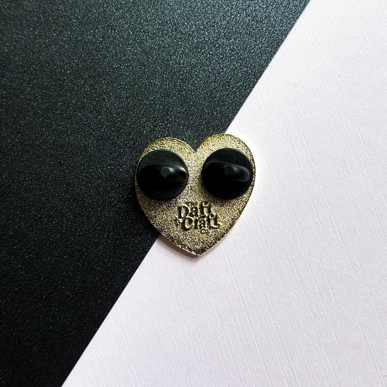 May include: A gold-colored heart-shaped enamel pin with two black pin backs. The pin has the text "Daft Craft Co." in a stylized font. The background is split diagonally, with black and white sections.