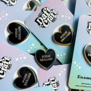 May include: Enamel heart-shaped pins with gold trim and the name "Dorian Havilliard" in gold lettering. The pins are on pastel-colored cards with the text "The Daft Craft Co." and star patterns. Some pins have a blue glitter border.