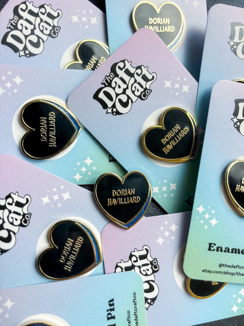 May include: Enamel heart-shaped pins with gold trim and the name "Dorian Havilliard" in gold lettering. The pins are on pastel-colored cards with the text "The Daft Craft Co." and star patterns. Some pins have a blue glitter border.