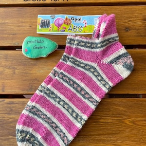 Könnte beinhalten: Ein handgestrickter Socken mit horizontalen Streifen in Pink, Grau und Creme. Der Socken wird zusammen mit einer Garnbanderole mit der Aufschrift Opal ABO und einem grünen Stein mit dem Text miniMata Socken gezeigt. Der Text Größe 40/41 ist sichtbar.