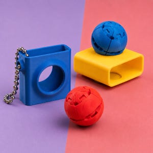 Könnte beinhalten: Ein blauer, rechteckiger Behälter mit einer kreisförmigen Öffnung und einer silbernen Kugelkette, neben einem gelben rechteckigen Block und einem roten Ball. Ein blauer Ball sitzt auf dem gelben Block. Die Objekte befinden sich auf einem lila und rosa Hintergrund.
