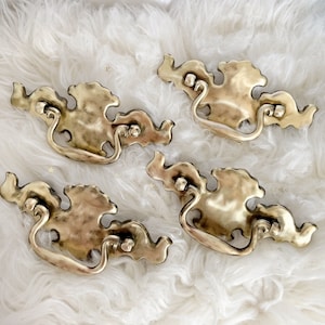 May include: Four ornate gold-toned drawer pulls with a decorative, slightly raised, floral design. The pulls have a loop handle and are set against a white fluffy background.