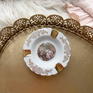 May include: White ceramic ashtray with gold trim and a floral design. The center features a vintage illustration of a couple in a garden setting.