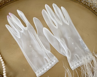 May include: A pair of sheer white gloves with delicate embroidered details and small circular cutouts. The gloves have a scalloped edge and are trimmed with white feathers. The gloves are displayed on a gold-colored tray.