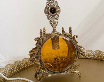 May include: An antique perfume bottle with a decorative filigree design. The bottle has a round, amber-colored glass body and is supported by an ornate, gold-toned metal base. A pearl necklace and a gold-colored tray are in the foreground.