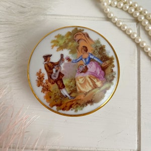 May include: A round porcelain trinket box with a gold rim. The lid features a painted scene of a couple in a garden setting. The man is playing a guitar and the woman is holding a basket of flowers. The scene is signed "Fragonard".