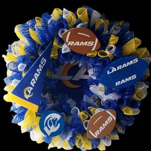 May include: A decorative wreath in blue, yellow, and white mesh, featuring football-shaped ornaments and a pennant, all bearing the "RAMS" logo. The wreath has a black background and the text "C&M Wreaths LLC".