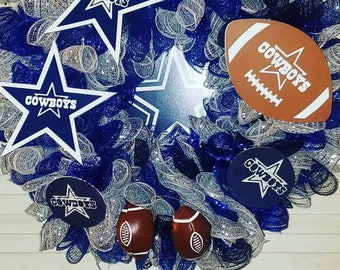 May include: A decorative wreath with blue, silver, and white mesh. It features star and American football-shaped ornaments with the word "Cowboys" and a star logo. A sports-themed wreath.