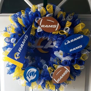 May include: A decorative wreath in blue, yellow, and white mesh, featuring football-shaped ornaments and a pennant with the text "RAMS." The wreath has the LA Rams logo in the center, perfect for sports fans.