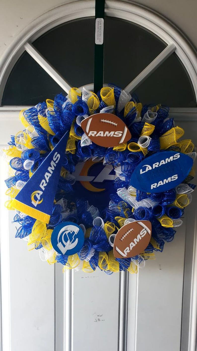 May include: A decorative wreath in blue, yellow, and white mesh, featuring football-shaped ornaments and a pennant with the text "RAMS." The wreath has the LA Rams logo in the center, perfect for sports fans.