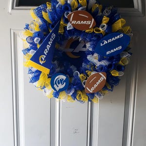 May include: A decorative wreath in blue, yellow, and white mesh, adorned with football-shaped ornaments and pennants. The wreath features the Los Angeles Rams logo and team name, perfect for displaying team spirit.