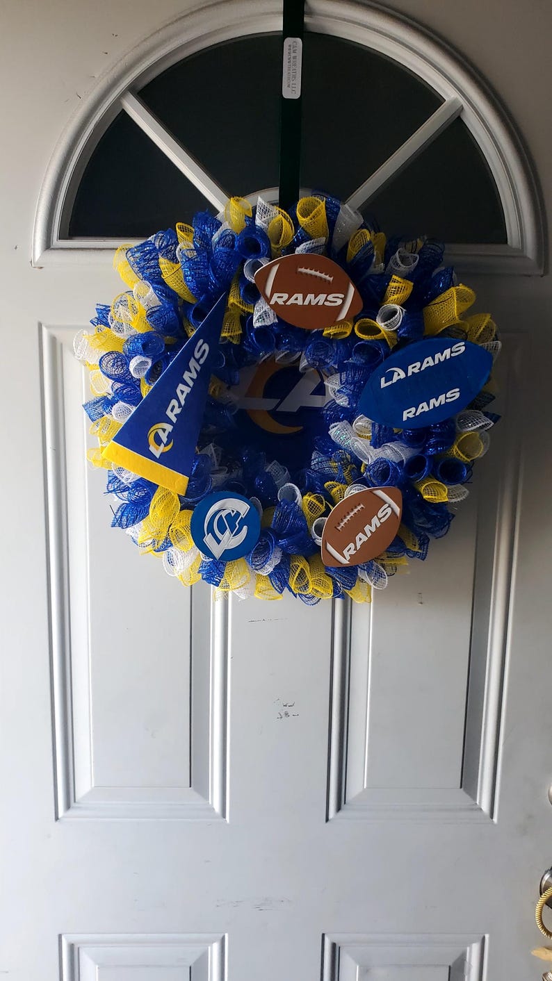 May include: A decorative wreath in blue, yellow, and white mesh, adorned with football-shaped ornaments and pennants. The wreath features the Los Angeles Rams logo and team name, perfect for displaying team spirit.