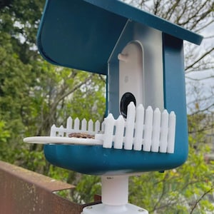 May include: A teal birdhouse with a white picket fence and a small perch. The birdhouse has a curved roof and is mounted on a white post. A small camera is visible inside the birdhouse.
