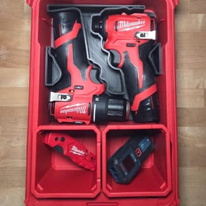 May include: A red Milwaukee tool case with a clear lid, containing red and black power tools. The case holds a drill, impact driver, a small utility knife, and a blue laser distance measurer.