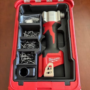 May include: A red and black Milwaukee power tool kit with a clear lid. The kit contains a red and silver power tool, nails, and other accessories. The Milwaukee logo is visible on the tool.