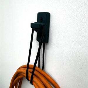 May include: A dark teal wall-mounted hook with a black cord holding an orange extension cord. The hook is rectangular with a curved lower section. The extension cord is coiled and secured with a black tie.