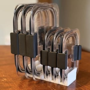 May include: A set of clear plastic food storage containers with black lids. The containers are stacked in a white organizer.