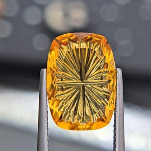 Könnte beinhalten: Ein leuchtend goldgelber Citrin-Edelstein mit rechteckigem Kissenschliff und einer detaillierten, sonnenstrahlenartigen Intaglio-Gravur auf der Oberfläche. Der transparente Stein wird von einer silberfarbenen Metallpinzette gehalten.