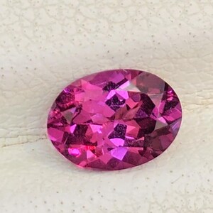 May include: A vibrant, oval-cut, pink gemstone. The gem is faceted, displaying multiple internal reflections and a brilliant shine. The stone is set against a white, textured surface, highlighting its color and clarity. This gemstone is suitable for jewelry.