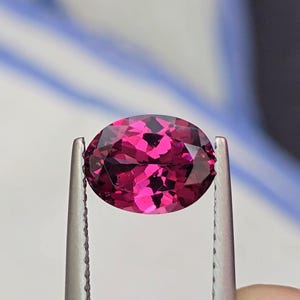 May include: A faceted, oval-shaped gemstone held by tweezers. The gem is a deep magenta color, with internal patterns visible. The tweezers are silver-colored, and the background is blurred, with hints of white and blue.