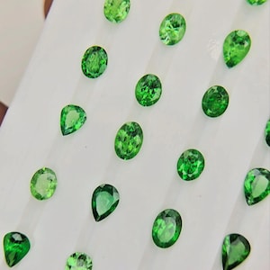 May include: A collection of loose, faceted green gemstones arranged in a grid pattern. The stones feature a mix of oval and pear shapes, each displaying a vibrant, translucent emerald-green hue with multiple facets that reflect light across the surface.