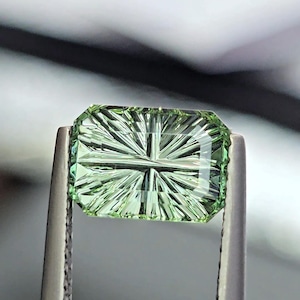 May include: A rectangular, light green gemstone featuring a precision-cut, sunburst-style faceting pattern that radiates from the center toward the edges. The stone is held securely by a pair of metal tweezers, showcasing its clarity and light reflection.