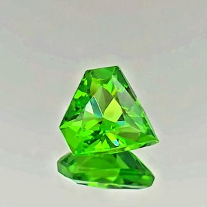 May include: A vibrant, lime green gemstone cut into a shield-like geometric shape with multiple facets that reflect light. The stone is positioned above its own clear reflection, showcasing a bright, translucent color and sharp, angular edges.