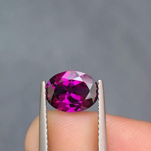 May include: A faceted, oval-shaped gemstone in a vibrant magenta hue. The gem is held by a pair of silver tweezers against a gray background. The stone's polished surfaces reflect light, showcasing its clarity and color saturation. A close-up view highlights the gem's brilliance.