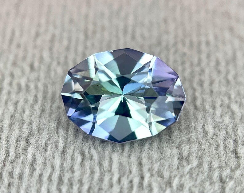 K&ouml;nnte beinhalten: Ein facettierter ovaler Edelstein in blaugr&uuml;ner Farbe. Der Edelstein hat einen hohen Glanz und ist vor einem wei&szlig;en Hintergrund platziert.