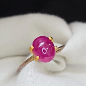 Peut inclure: Une bague en or ornée d'une grosse pierre précieuse rose vif de forme ovale. La pierre est maintenue par quatre griffes en or. Le corps de la bague est fin et de couleur or. Le fond est un tissu blanc doux.