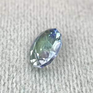 K&ouml;nnte beinhalten: Ein facettierter ovaler Edelstein mit einem blaugr&uuml;nen Farbverlauf. Der Edelstein ist vor einem wei&szlig;en, flauschigen Hintergrund platziert.