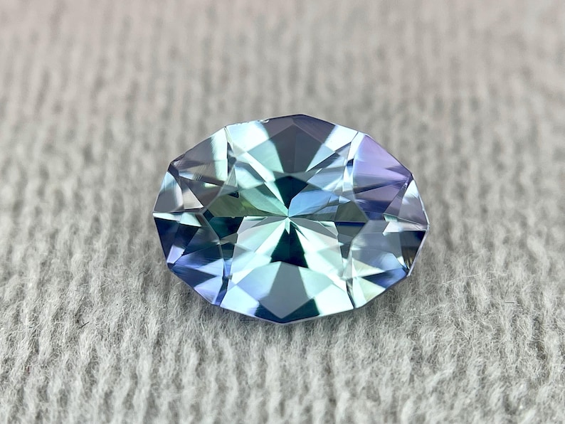 May include: A faceted oval-shaped gemstone with a blue-green hue. The gem has a star-like pattern in the center.