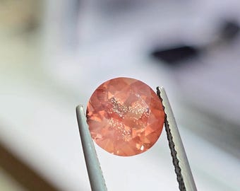 May include: A round, faceted gemstone with a peach-orange hue, held by tweezers. The gem has a sparkling, glittery appearance. The background is blurred, suggesting a studio setting. This gemstone is likely for jewelry making.