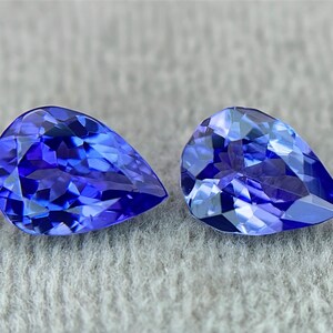 May include: Two pear-shaped blue gemstones, likely Tanzanite, are displayed on a gray textured surface. The gemstones are faceted and have a deep blue color with a slight purple hue.