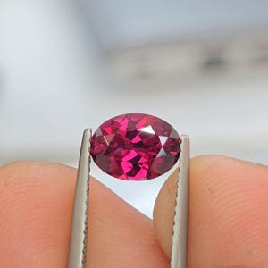 May include: A faceted, oval-shaped gemstone held by tweezers. The gem is a deep, vibrant red color, with multiple facets catching the light. The background is blurred, emphasizing the gem's brilliance.