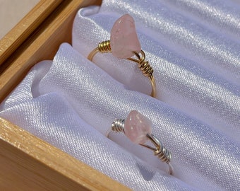 May include: Two rings with pink rose quartz stones wrapped in gold and silver wire. The rings are displayed in a wooden box lined with white satin.