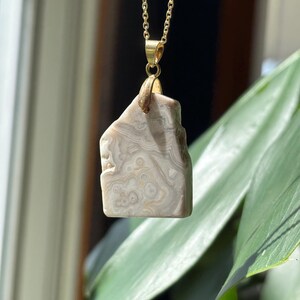 Puede incluir: Collar con cadena dorada y colgante de piedra clara. La piedra presenta un patrón único de remolinos y líneas en tonos beige y gris. El colgante es rectangular con bordes redondeados.