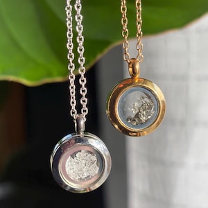 May include: Two necklaces with round pendants, one silver-toned and one gold-toned. Each pendant contains a small, clear glass orb filled with a cluster of shiny, irregular-shaped pieces. The necklaces hang against a blurred green background.