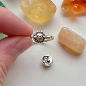 Op de afbeelding: Een zilveren ring met een zonontwerp. De ring ligt op een wit oppervlak met verschillende kleine stenen.