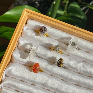 May include: A set of six rings featuring different natural stones wrapped in gold wire. The stones include amethyst, clear quartz, tiger's eye, carnelian, and two other stones.