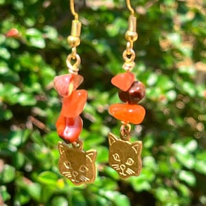 May include: Gold-colored dangle earrings featuring a cat face charm. Each earring has a stack of orange and brown gemstone beads. The earrings hang from gold-colored ear wires. The background is a blurred view of green foliage.