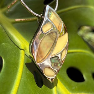 May include: A silver-toned pendant necklace with a unique, abstract design. The pendant features a polished silver frame with inlaid sections of cream and iridescent shell-like material. The necklace is displayed against a green leaf background.