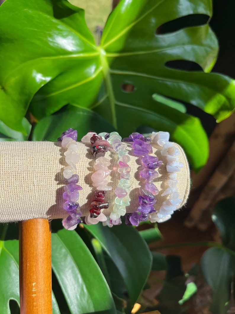 K&ouml;nnte beinhalten: Vier Edelstein-Armb&auml;nder in verschiedenen Farben, darunter Amethyst, Rosenquarz, gr&uuml;ner Aventurin und blauer Achat. Die Armb&auml;nder sind auf einem Holzst&auml;nder gestapelt.
